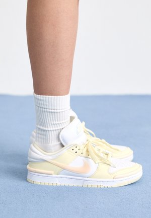 Fod iført hvide ribstrikkede sokker og hvide og lysegule Nike-sneakers, står på en lyseblå tekstureret overflade.