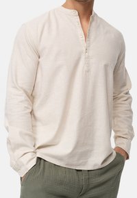 Mężczyzna w beżowej, lnianej koszuli z długim rękawem i kołnierzykiem band, w oliwkowych, teksturowanych spodniach, z rękami w kieszeniach, skróconych do szyi.