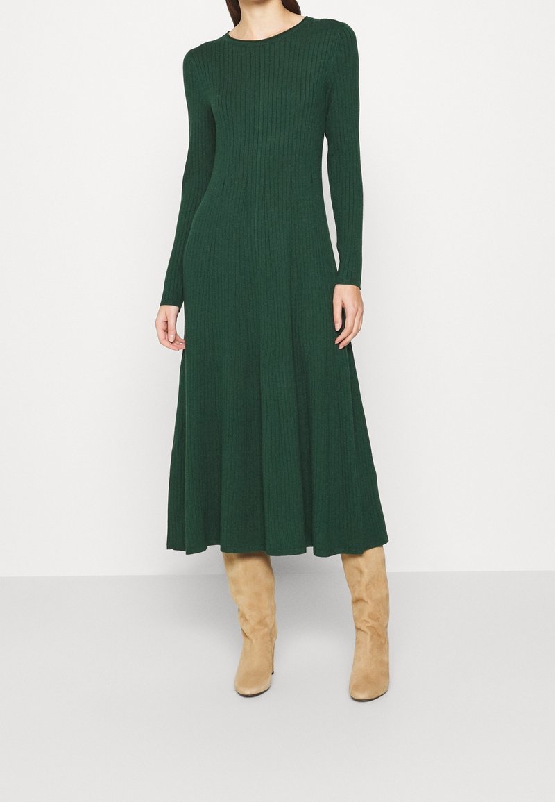 Robe verte à manches longues, côtelée, avec une jupe évasée. Le mannequin porte des bottes beige montantes jusqu'au genou, mettant en valeur la silhouette fluide de la robe.