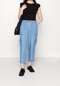 Top nero con maniche arricciate, pantaloni cargo in denim azzurro chiaro con vita regolabile e tasche laterali, abbinati a scarpe slip-on nere.