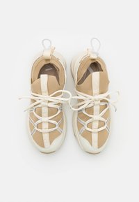 Zapatillas deportivas beige con un upper de tejido texturizado, detalles en crema, cordones blancos y lengüetas de tirón, que presentan una suela ligeramente gruesa y costuras decorativas.