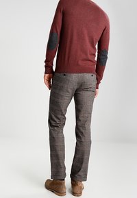 Burgundowy sweter z dzianiny z szarymi łaty na łokciach, zestawiony z szarymi spodniami w kratę. Model pokazuje widok z tyłu, nosząc brązowe zamszowe buty.