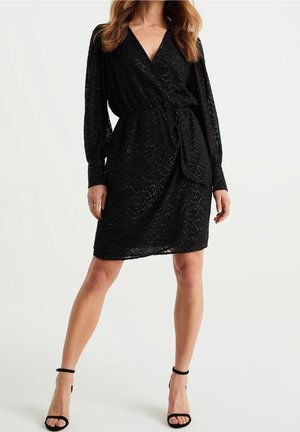 Femme portant une robe portefeuille noire mi-longue avec des manches longues transparentes et des escarpins noirs à bride à la cheville, debout sur un fond blanc.