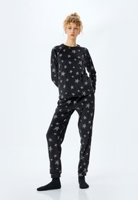 Jonge vrouw staat, gekleed in een zwart bijpassend top met sterrenpatroon en een broek, met zwarte sokken tegen een plain witte achtergrond.