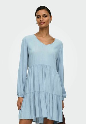 Femme portant une robe bleu clair à étages avec manches longues et col en V, tenant un sac brun foncé sur un fond uni.