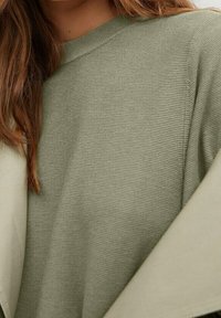 Lichtgroene gebreide trui met een ronde hals en geribbelde textuur, gedragen onder een beige jas of coat, gedeeltelijk zichtbaar in het kader.