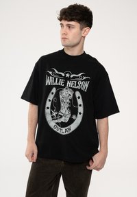 Black oversized T-shirt featuring a graphic print of a cowboy boot encircled by stars and the text "WILLIE NELSON OUTLAW" in contrasting colors.