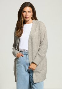 Donna con lunghi capelli castani che indossa un cardigan a maglia beige, una maglietta bianca e jeans a vita alta blu chiaro, in piedi con una mano in tasca.
