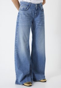 Persona que lleva vaqueros de tiro alto y pierna ancha de color azul con detalles frontales desgastados, combinados con zapatos dorados brillantes de punta cuadrada.