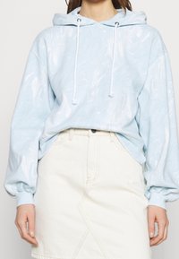 Lyseblå hættetrøje med marmoreret tekstur, der har snor i hætten og oversized ærmer, parret med en hvid denimnederdel.
