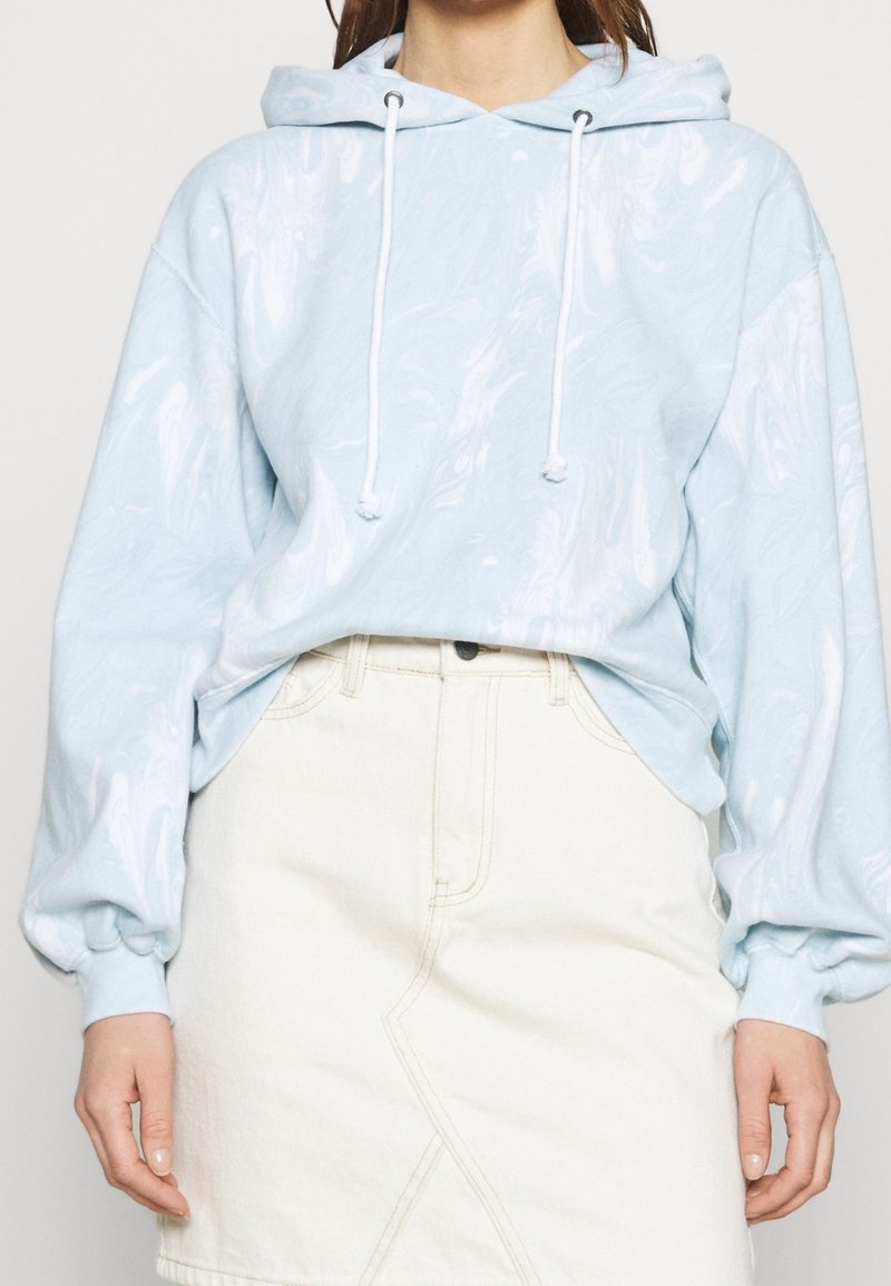 Lyseblå hættetrøje med marmoreret tekstur, der har snor i hætten og oversized ærmer, parret med en hvid denimnederdel.