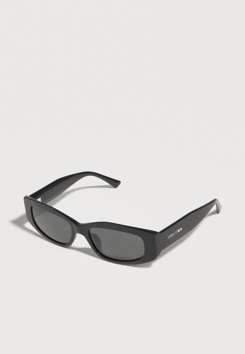 Μαύρα ορθογώνια γυαλιά ηλίου με σκούρους φακούς και χοντρά πλαίσια, γωνιακά για να φαίνεται ο αριστερός βραχίονας με το λευκό λογότυπο "Jimmy Choo".