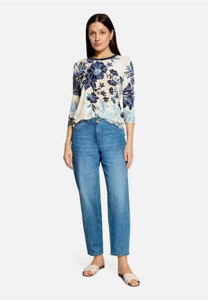Frau steht und trägt ein cremefarbenes Oberteil mit blauem Blumenmuster, blaue hoch taillierte Jeans und beige Slip-on-Sandalen auf weißem Hintergrund.