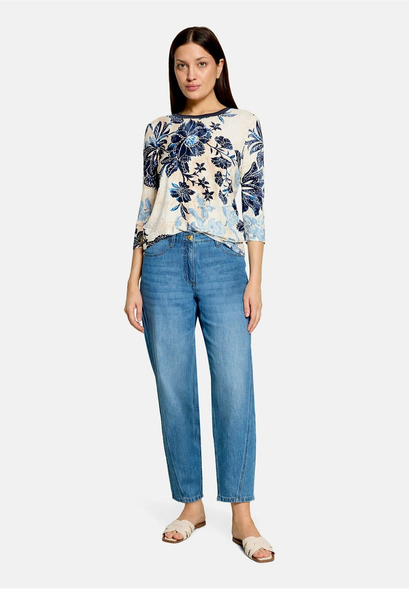 Femme debout portant un haut crème à imprimé floral bleu, un jean bleu taille haute et des sandales beiges à enfiler, sur fond blanc.