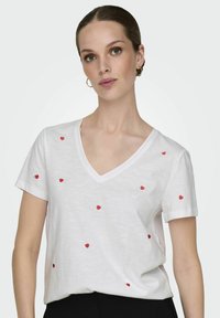 T-shirt blanc à manches courtes et col en V orné de cœurs rouges brodés éparpillés. Tissu doux avec une coupe décontractée, associé à un pantalon noir.