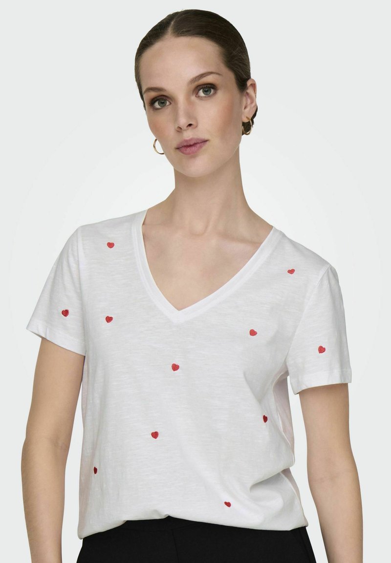 T-shirt blanc à manches courtes et col en V orné de cœurs rouges brodés éparpillés. Tissu doux avec une coupe décontractée, associé à un pantalon noir.