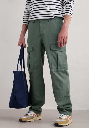 Cargo pants in olive green cotton, with side pockets and a straight fit. Worn with a striped long-sleeve shirt and beige sneakers, carrying a navy bag.