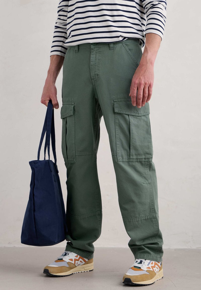 Cargo-byxor i olivgrön bomull, med sidofickor och rak passform. Bärs med en randig långärmad skjorta och beige sneakers, hållande en marinblå väska.