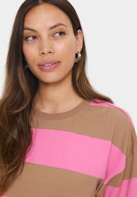 Femme aux longs cheveux bruns portant des boucles d'oreilles en argent et une chemise rayée marron et rose, regardant légèrement sur le côté.