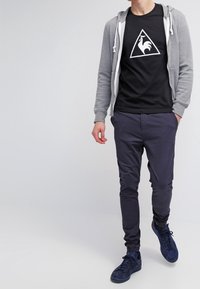 Černé tričko s bílým trojúhelníkovým logem, šedá zipová mikina, námořnické kalhoty a modré tenisky. Jednoduchý design s volným střihem.