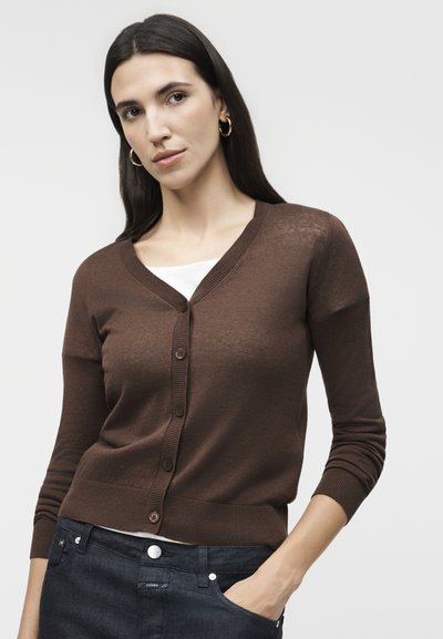 Γυναίκα με μακριά σκούρα μαλλιά που φορά χρυσούς κρίκους στα αυτιά, καφέ κουμπωτό ζακέτα πάνω από λευκό τοπ και σκούρο τζιν, με το ένα χέρι στην τσέπη.