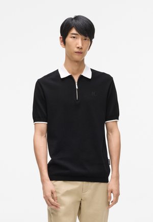 Fekete kötött pólóing fehér gallérral és cipzárral. Rövidujjú, az mellkason hímzett logóval rendelkezik. Sima textúra.