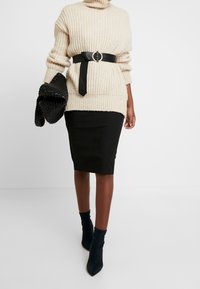 Pull beige côtelé à col montant, taille ceinturée noire, jupe crayon noire et bottines noires à bouts pointus. Pochette tissée noire.