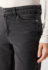 Jean en denim noir avec une coupe classique, poches avant et bouton en métal. Le tissu a une légère texture avec un minimum de décoloration.