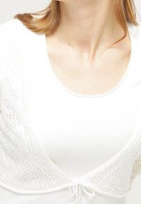 Vitt stickat linne med spetsmönster. Har en rund halsringning och ett knytband fram, vilket framhäver textur och design.