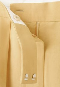 Pantalon beige avec fermeture par bouton et crochet-barre montrant les détails de la ceinture intérieure et de la fixation.