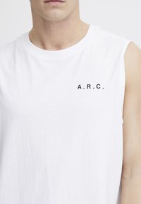 Valge ilma varrukateta puuvillane tipp, millel on ümmargune kaelus ja must print "A.R.C." logo vasakul rinnal. Sile tekstuur ja mugav lõige.