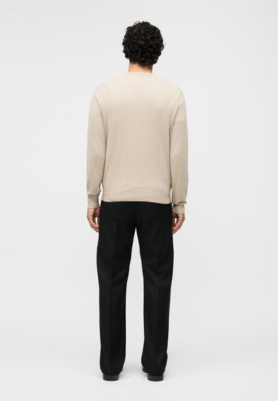 Άνδρας με σγουρά μαλλιά, φορώντας μπεζ πουλόβερ με μακριά μανίκια και μαύρο παντελόνι με ίσια γραμμή, στέκεται γυρισμένος με την πλάτη προς το μονόχρωμο φόντο.