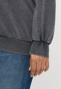 Sweatshirt cinza com punhos e barra canelados, apresentando um corte descontraído e uma textura suave. Mão repousada ao lado, combinado com jeans.