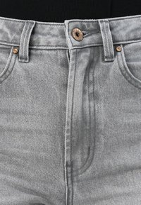 Gros plan sur la taille d’un jean en denim gris avec bouton en métal, passants de ceinture et poches avant sur fond noir.
