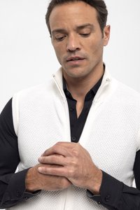 Chaleco de punto blanco con cremallera sobre una camisa negra con cuello, mostrando un patrón texturizado y un detalle de cremallera visible. Las manos están entrelazadas al frente.