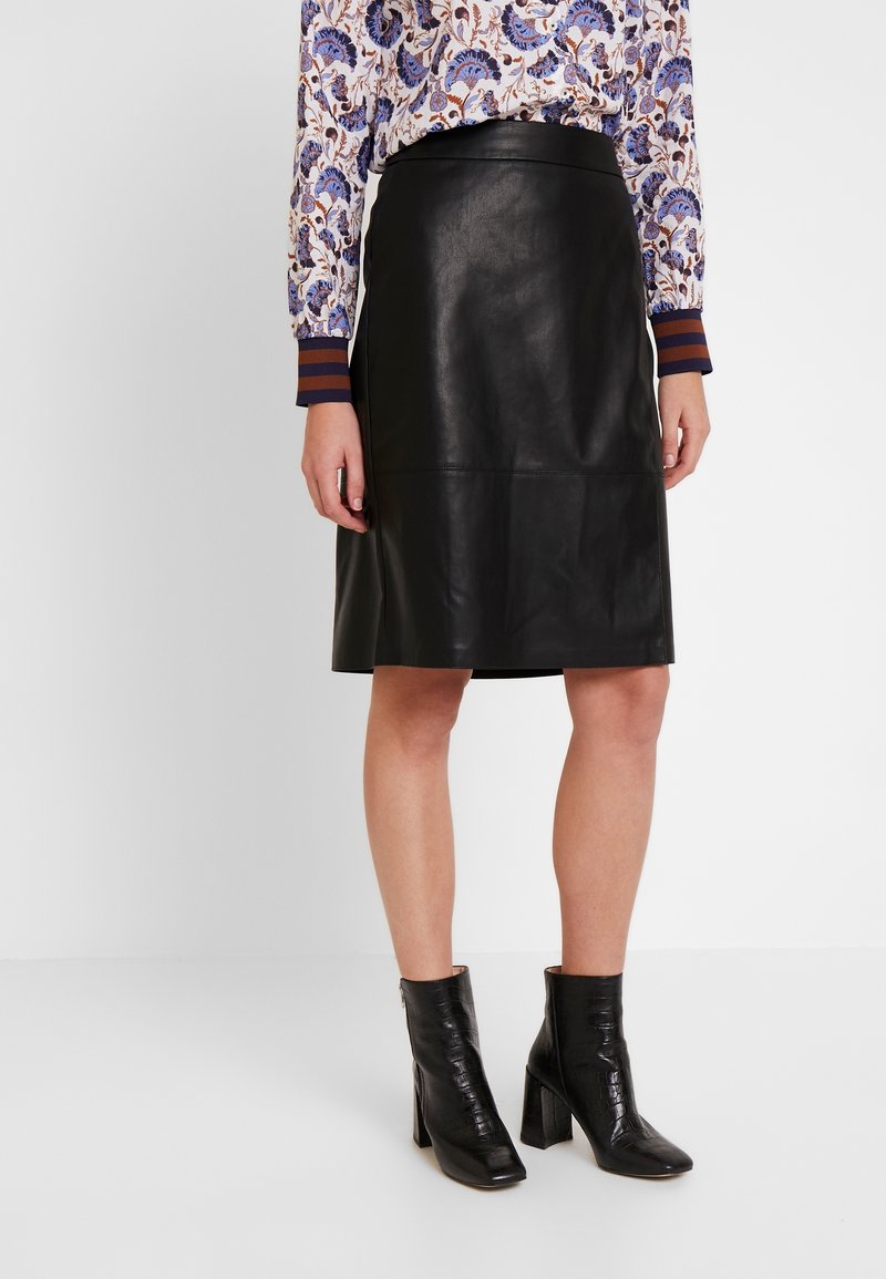 Femme portant une jupe en cuir noire longueur genou, un chemisier à fleurs avec des poignets rayés et des bottines noires à talons, debout sur un fond blanc.