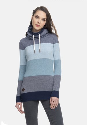 Kobieta ubrana w niebieski sweter w paski z wysokim kołnierzem typu komin i białymi sznurkami, w połączeniu z ciemnoszarymi spodniami.