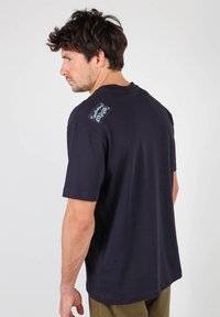 T-shirt à manches courtes bleu marine en coton, avec un patch sur l'épaule gauche portant un design de logo. Texture lisse et coupe classique.