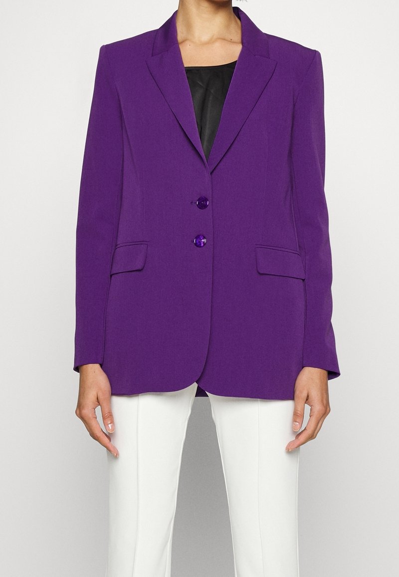 Femme portant un blazer violet sur mesure avec deux boutons par-dessus un top noir et un pantalon blanc, debout devant un fond uni.