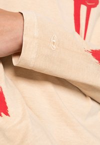 Beige pitkähihaine paita, jossa on teksturoitu kangas. Ominaisuuksiin kuuluu punainen maalisuihkusuunnittelu ja pieni valkoinen logo-embroidointi hihansuun kohdalla.