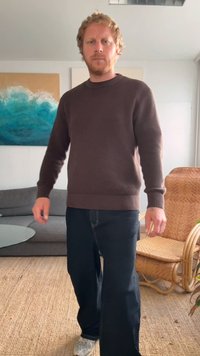 Maglione marrone con texture e orlo a costine, abbinato a jeans a gamba larga scuri. Presentato in un interno moderno con una sedia intrecciata e un tavolo di vetro.