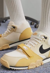 Tenisky Nike s kombinací bílých, krémových a žlutých materiálů, s černými akcenty, se zapínáním na pásku na přední straně a texturovanou podrážkou.