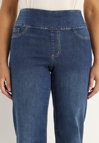 Vrouw in hoog-was blauwe denim jeans met zichtbare stiksels, voorzakken en een witte top erin gestoken.