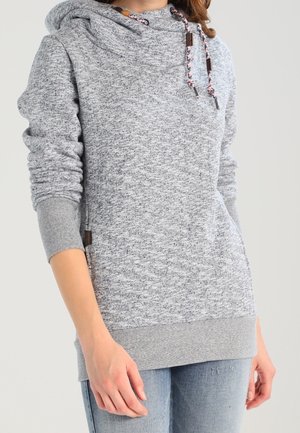 Osoba nosící světle šedou melírovanou mikinu s kapucí a stahovacími šňůrkami a vybledlé modré džíny, stojící před jednoduchým pozadím.