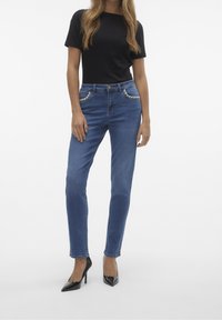 Jeans de mezclilla azul con corte recto, que cuentan con bolsillos acentuados en plata y una cintura alta, combinados con una blusa negra ajustada de mangas cortas y tacones negros.