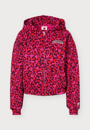 Un mannequin se tient devant un fond uni, portant un sweat à capuche zippé rose vif à imprimé léopard, un pantalon assorti à jambes larges et des chaussures noires.