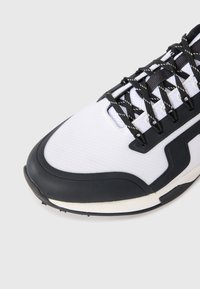 Balti un melni sporta apavi ar melnām auklām, atstarotāju svītrām, sietveida augšdaļu un gumijas zoli uz vienkārša fona.