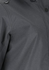 Mørkegråt jakke stof med synlige syninger, mærketag nær skulderen og en rund sort trykknap foran.