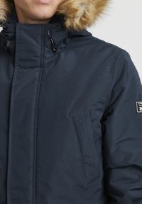 Navy blå pufferjakke med hette i pelskant, med en vertikal ribbet lomme og sorte detaljer. Glatt tekstur og isolert design.