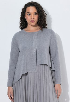 Vrouw met krullend haar die een grijze asymmetrische trui met lange mouwen draagt over een geplooide rok, staande tegen een effen witte achtergrond.